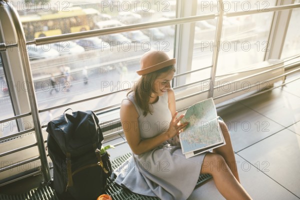 Waiting, delayed transport in the terminal of the airport or train station. Young caucasian woman in dress and hat sits on tourist rug with backpack near window to room and looks into tourist map