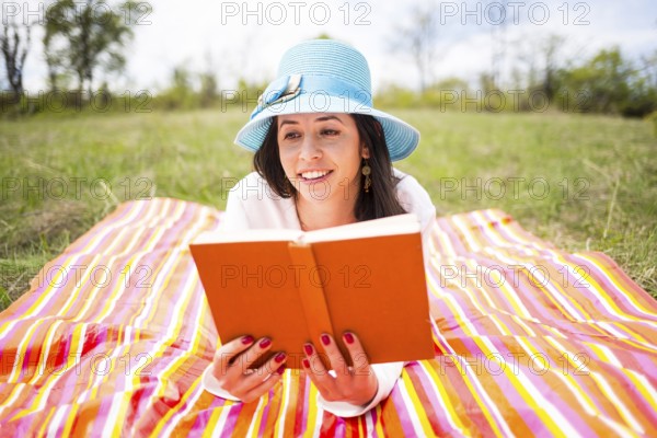 Attractive smiling young woman dressed casually reads orange old book lying down on a colourful blanket in the park. Traditional reading