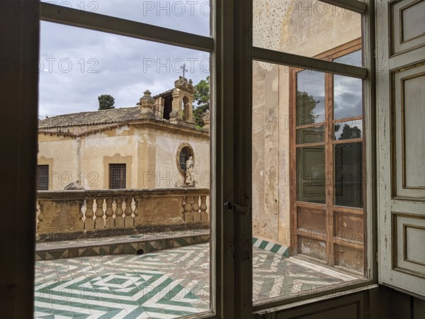 A window view of a historic terrace with patterned floor tiles and vintage architectural elements, Villa Palagonia, known as the ‘villa of monsters', is an 18th-century walled building located in Bagheria, Sicily