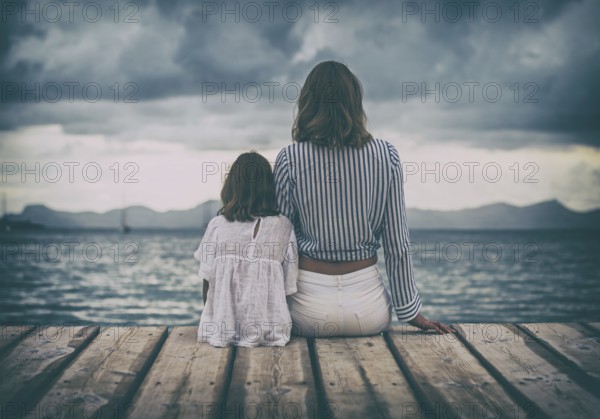 Woman and little girl sitting on the wooden pier near the sea