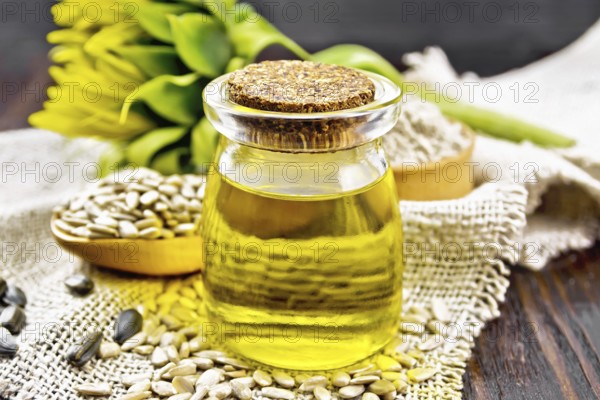 Sunflower oil in glass jar, spoon with peeled seeds and flour in bowl on burlap, a flower on wooden board background