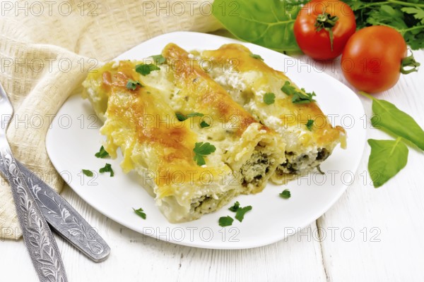 Cannelloni stuffed with cottage cheese and spinach with Bechamel sauce in a plate, towel, tomatoes and parsley on the background of light wooden board