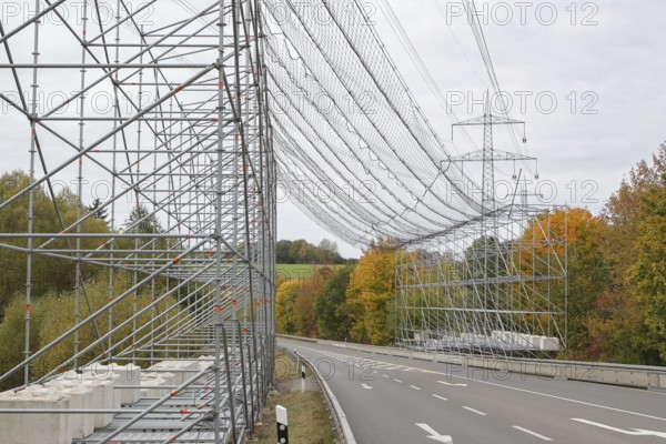 A scaffold with steel net as protection over a road, safety measure due to work on an overhead line and electricity pylons, in the background trees in autumn