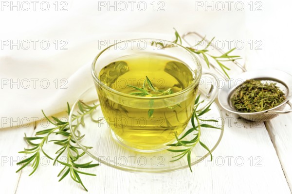 Rosemary herbal tea in a cup, a strainer with dry herb and a linen napkin on the background of a light wooden board