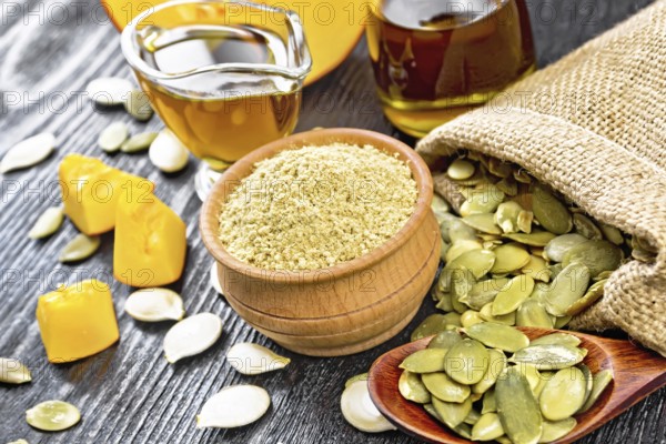 Pumpkin flour in a bowl, oil in a sauceboat and a jar, seeds in bag, on table and in spoon, slices of vegetable on dark wooden board background