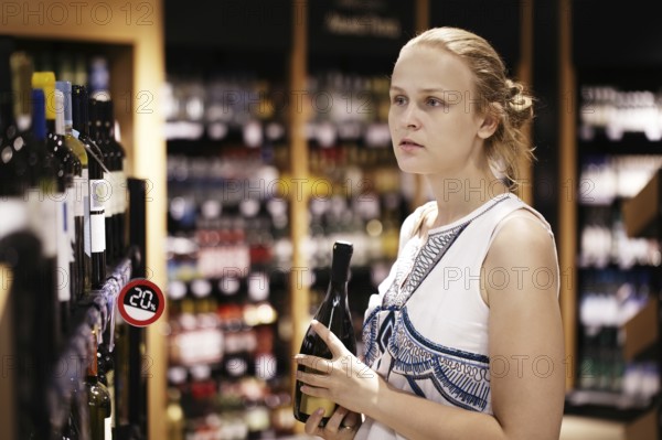 Woman shopping for wine or other alcohol in a bottle store standing in front of shelves full of bottles with a serious expression as she tries to make up her mind