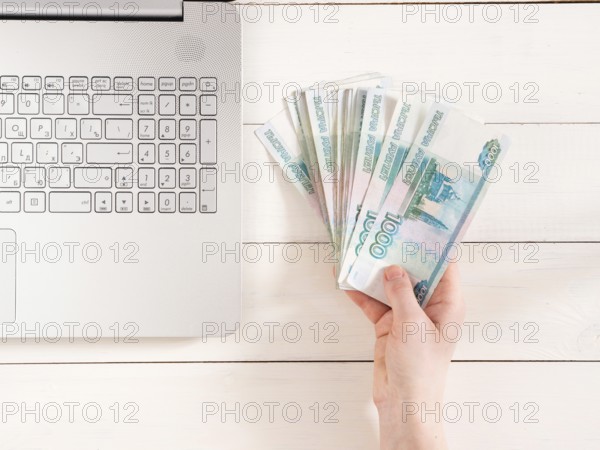 Hand holding russian rubles near laptop on white wooden background. Top view or flat lay. Finance and earning concept