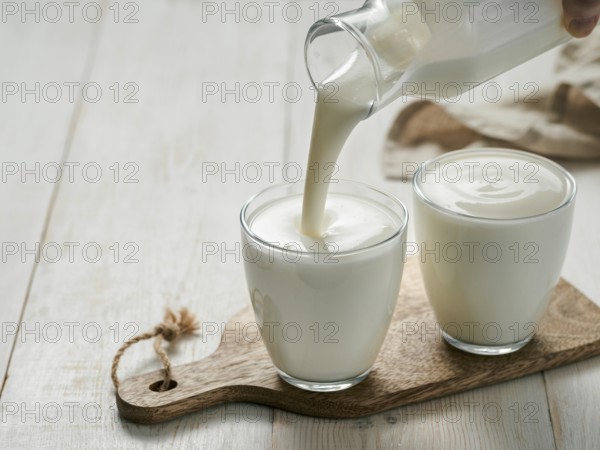 Pouring homemade kefir, buttermilk or yogurt with probiotics. Yogurt flowing from glass bottle on white wooden background. Probiotic cold fermented dairy drink. Trendy food and drink. Copy space left
