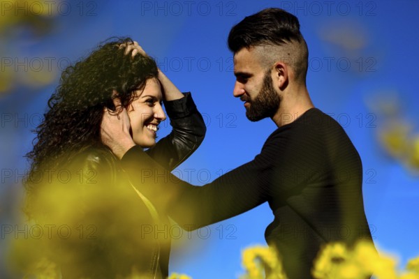 Portrait of a young smiling couple where hair man caresses his girlfriend's face before kissing blue background and yellow flowers out of focus in the foreground