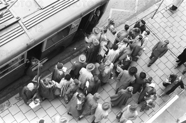 Bus stop in Stockholm in 1953. People are waiting for the bus at the bus stop. Bus 32 travelling on the Fleminggatan-Fridhemsplan line has stopped and people have started to get on the bus. The bus was powered by electricity and was called a trolleybus, it draw it's power from overhead wires using spring-loaded trolley poles. Stockholm 1953, Kristoffersson ref 1-11