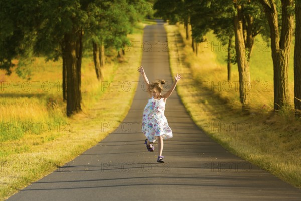 A little girl in a summer dress runs along a street in the light of a summer evening, jumping and gesticulating joyfully. A picture of perfect happiness! Little girl running along a road jumping throws her arms in the air in a joyous gesture. Warm sunlight of a summer evening
