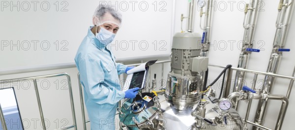 Widescreen picture, scientist in blue laboratory suit and gas mask stand in clean room and check graphs in tablet near big steel tank