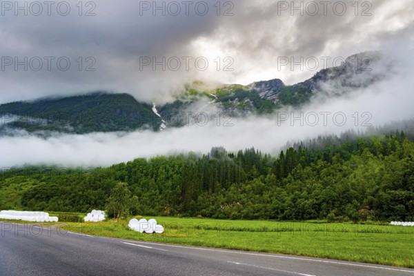 Wide asphalt road crosses the valley. Jostedalsbreen Park. Valley surrounded by high forested mountains. Heavy clouds covered the sky. Cold summer in Norway