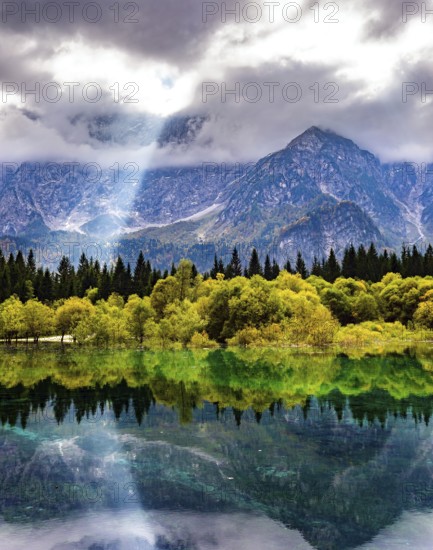 The sun's rays break through the clouds. The lake Fusine in Northern Italy. Mountains covered in mist. The smooth surface of the water reflects forest and mountains. Gorgeous reflections