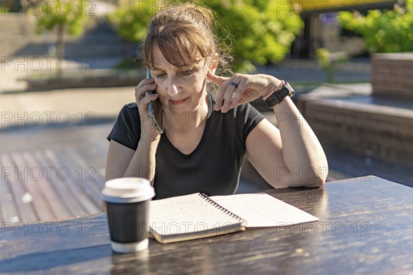 Woman over 50 reviewing notes while on a call with coffee outdoors