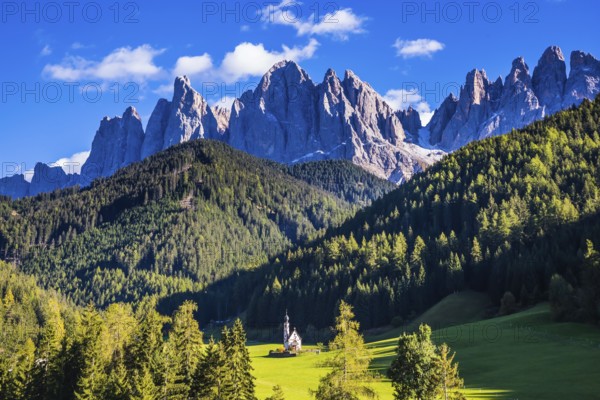 Dolomites, Tirol. Sunny autumn day. Rocky peaks and forested mountains surrounded by green Alpine meadows. The symbol of the valley Val di Funes - church of Santa Maddalena