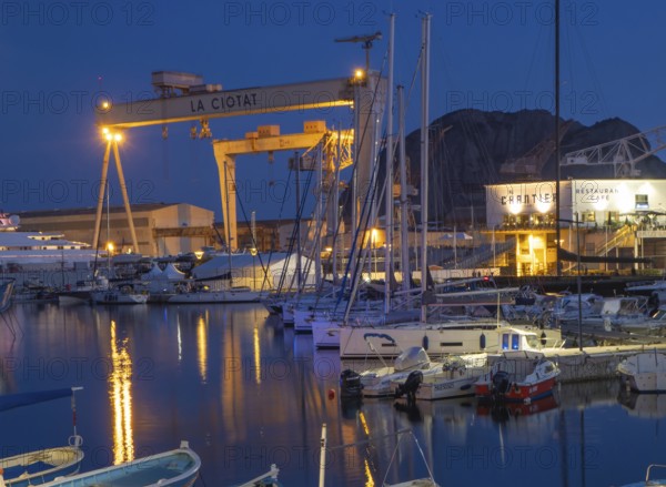 The famous harbour of La Ciotat, France, with fishing and sailing boats, in front of the industrial area with the illuminated dockside crane. Situation in the evening during blue hour