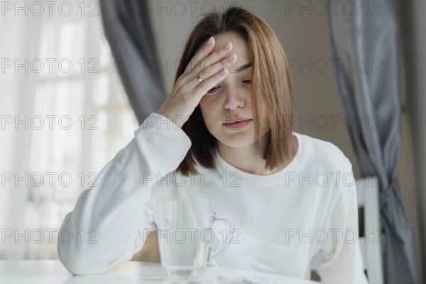 Woman holding her forehead looking fatigued, sitting indoors with a glass of water, suggesting a headache or stress