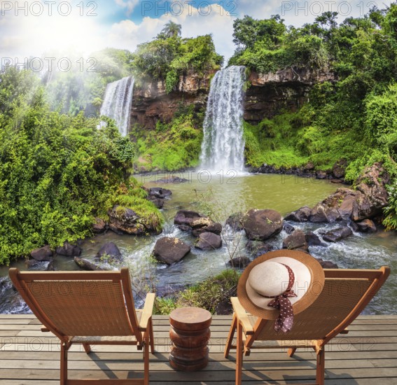 Two waterfalls from Iguazu Falls fall into a small quiet pond. The concept of ecological tourism. Two wooden chaise longue stand near the waterfalls. On the armchair hangs an female straw hat