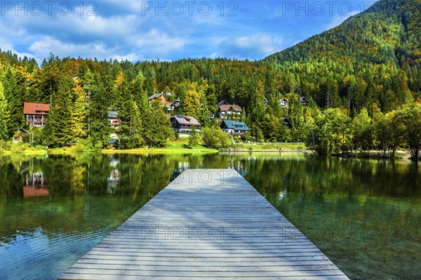 Travel to fabulous Slovenia. Lake Jasna in the Eastern Julian Alps. Wooden pier. The mountains are overgrown with dense mixed forest. On the banks there are lovely houses with red roofs
