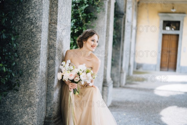 Smiling bride in a dress with a bouquet of pink flowers sat down near a pillar in a vaulted room. Lake Como.