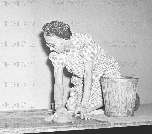 Elderly cleaning lady is on her knees scrubbing floorboards dressed in a classic cleaning coat and huckle. Despite the fact that the work is boring and laborious, she can be seen to have a positive expression on her face. Sweden 1947. Kristoffersson AD15-12