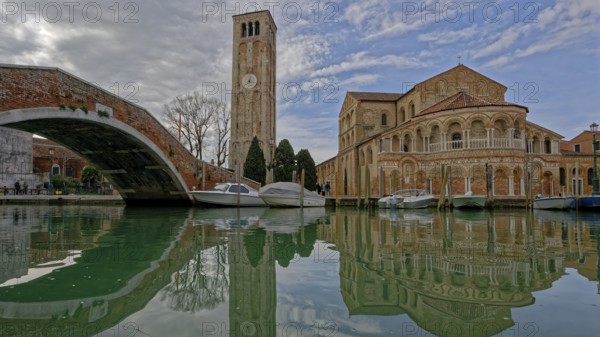 Scenic canal with boats reflecting a historic church and tall bell tower under a cloudy sky, Ponte san Donato and Basilica dei Santi Maria e Donato, Murano, Veneto, Italy
