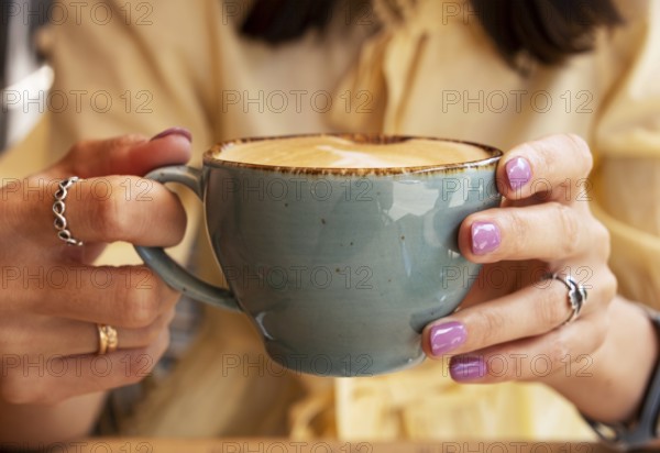 Woman in yellow clothes holding blue cup of cappuccino with both hands, close-up