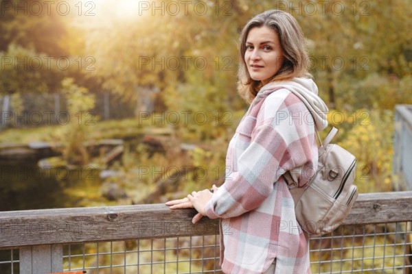 Portrait of cute young woman in casual wear in autumn, standing on bridge against background of an autumn Park and river. Pretty female walking in Park in golden fall. Copy space. smiling girl in the park standing on wooden bridge and looking at the camera in autumn season