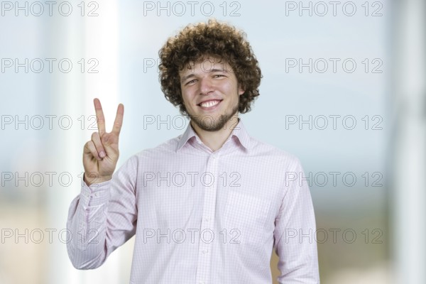 Portrait of a happy young man with curly hair shows victory gesture sign. Indoor window in the background