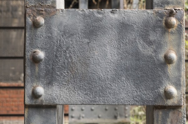 Close-up of a weathered gray metal plate attached to a larger metal structure with rivets, showing signs of rust and wear