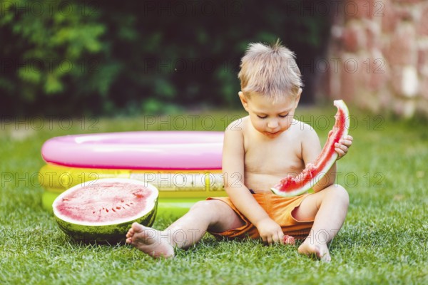 Theme summer seasonal food juicy watermelon berry. small funny Caucasian boy child two years sitting backyard green grass, lawn and eating piece watermelon holding hands. Naked in shorts and barefoot