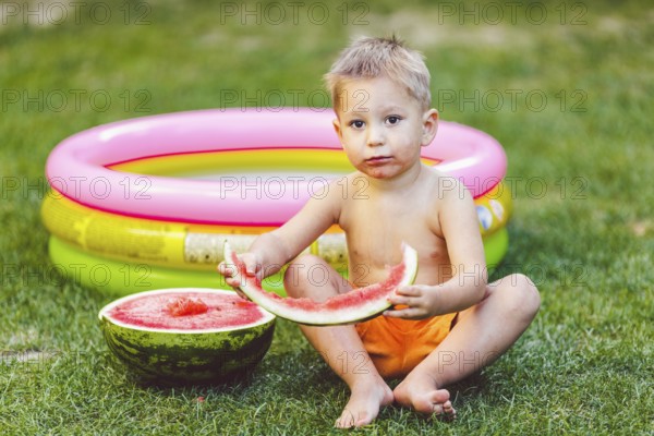 Theme summer seasonal food juicy watermelon berry. small funny Caucasian boy child two years sitting backyard green grass, lawn and eating piece watermelon holding hands. Naked in shorts and barefoot