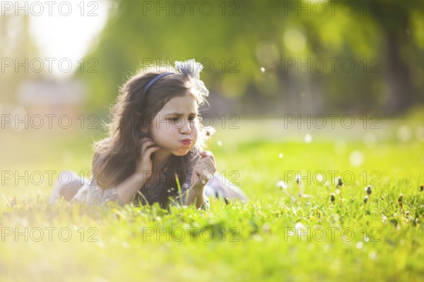 The little girl lays on a grass and blowing dandelion on the summer day