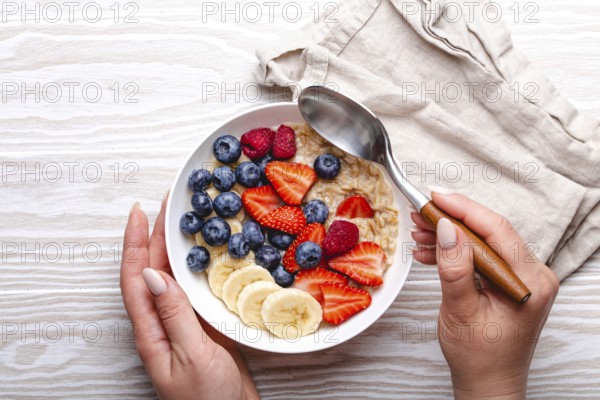 Top view of female hands holding bowl with oatmeal porridge with fruit and berries and spoon on white wooden background table top view, woman eating homemade healthy breakfast cereal, food photography