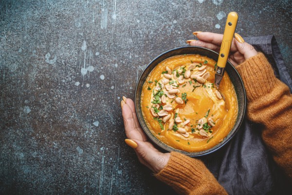 Female hands in yellow knitted sweater holding a bowl with pumpkin cream soup on dark stone background with spoon decorated with peanuts, top view. Autumn cozy dinner concept, space for text, food photography