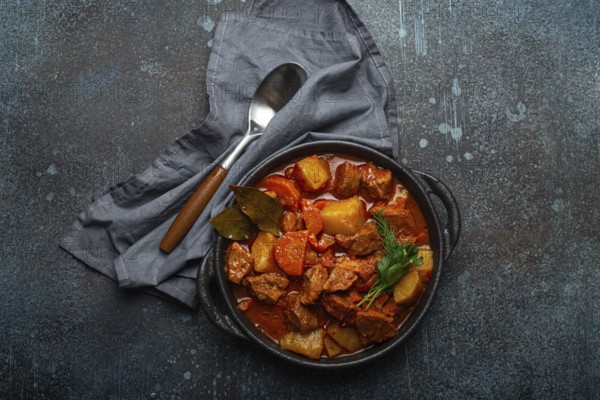 Beef meat stew with potatoes, carrot and delicious gravy in black casserole pot with bay leaves and fresh green herbs with spoon on black dark rustic concrete background from above, food photography