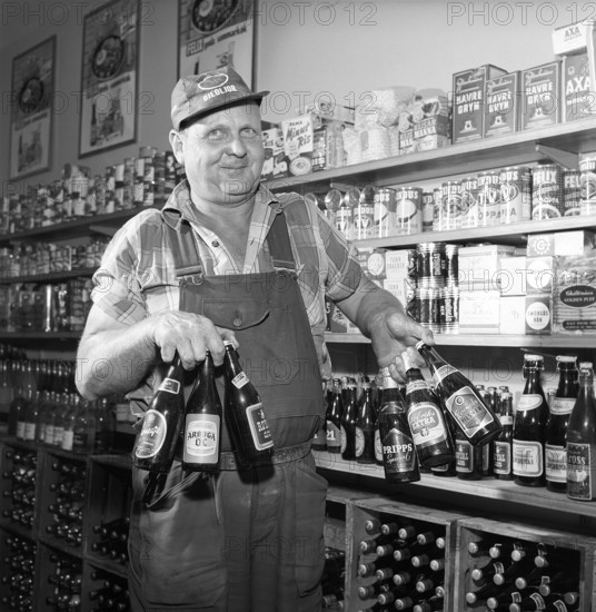 Large selection of beer and lager in 1960. A man with a good grip manages to hold six beer bottles in both hands at the same time. Famous beer brands at the time such as Carnegie, Arboga beer, Stockholms bryggerier, Pripps Special, St Eriks Extra and Risingsbo pilsner. He is standing in his work clothes with a cap on that has the gasoline chain OK biloljor in the text. In the background you can see that he is standing in a small grocery store with all sorts of different foods visible on the shelves. Sweden 1960. Conard ref 4269