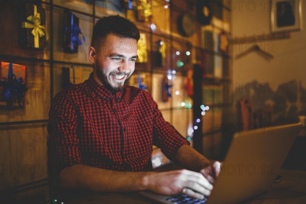 Theme training and computers. A young man with a beard and in a shirt uses a laptop, prints on the keyboard in a coffee shop at a wooden table in the evening. Christmas decor and hang a garland