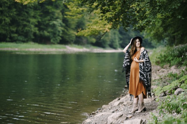 Pregnant woman stands on a rocky lake shore in the forest.