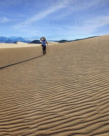 USA. Mesquite Flat Sand Dunes, Death Valley, California. Woman with a camera and a tripod goes among the dunes. Light sand waves from the desert wind. Concept of active and photo tourism