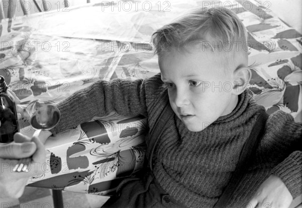 A boy is sick and won't take his medicine. His mother holds out a spoon and waits for him to gape and take it, but he looks very suspicous on the spoon. An everyday life event that today would look the same, the medicine sometimes does not taste so good. Sweden 1956. Conard ref 3157