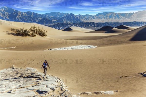 Elderly woman with a camera and a tripod goes among the dunes. Light sand waves from the desert wind. Mesquite Flat Sand Dunes, Death Valley, California. Concept of active and photo tourism