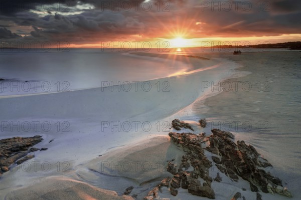 Picturesque La Pelosa beach in Sardinia at sunrise with a sunburst and rocks in the foreground