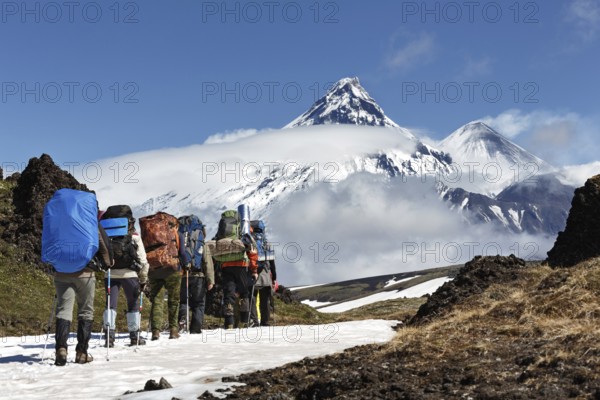 Group of hikers climbing in Kamchatka mountain on background of Klyuchevskaya group of volcanoes: Kamen Volcano, active Klyuchevskoy Volcano and active Bezymianny Volcano on sunny day. Russian Federation, Far East, Kamchatka Peninsula