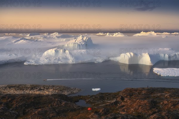 Large glaciers in the bay by the rocky shore in the light of the setting sun with fog over the water with orange tourist tent