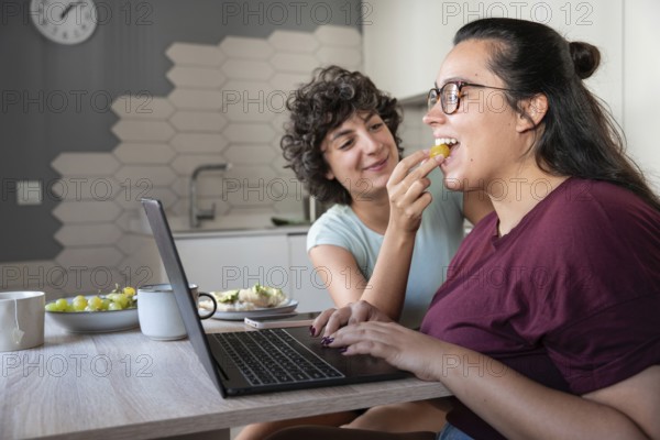 A happy lesbian couple enjoys feeding each other while working. Their laughter, casual attire, and homey setting convey warmth and love