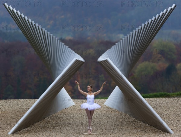 Ballerina in white ballet costume in elegant pose in front of modern sculpture in autumnal landscape, Emmendingen, Baden-Württemberg, Germany