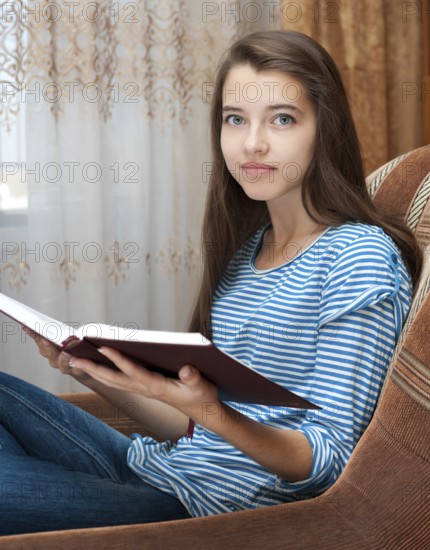 A young caucasian teenage girl reads a book sitting in an armchair