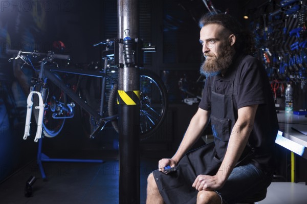 Portrait of small business owner of young man with beard. Guy bicycle mechanic workshop worker sitting with tool in his hand in a working black clothes in an apron in the background of a bicycle shop
